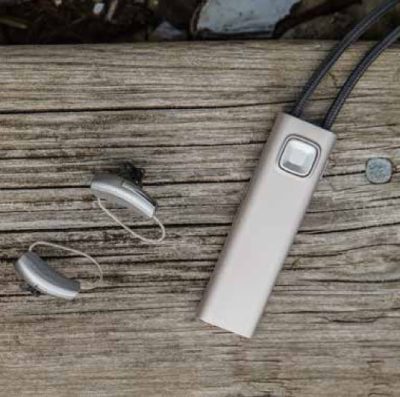 Two hearing aids and a rectangular beige Widex COM-DEX device with wires are placed on an outdoor weathered wooden surface. A small coin lies nearby.