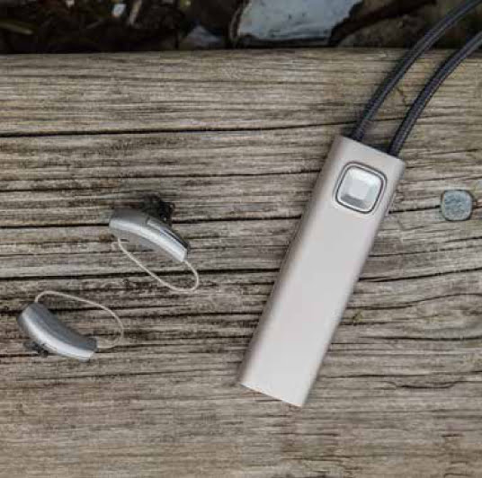 Two hearing aids and a rectangular beige Widex COM-DEX device with wires are placed on an outdoor weathered wooden surface. A small coin lies nearby.